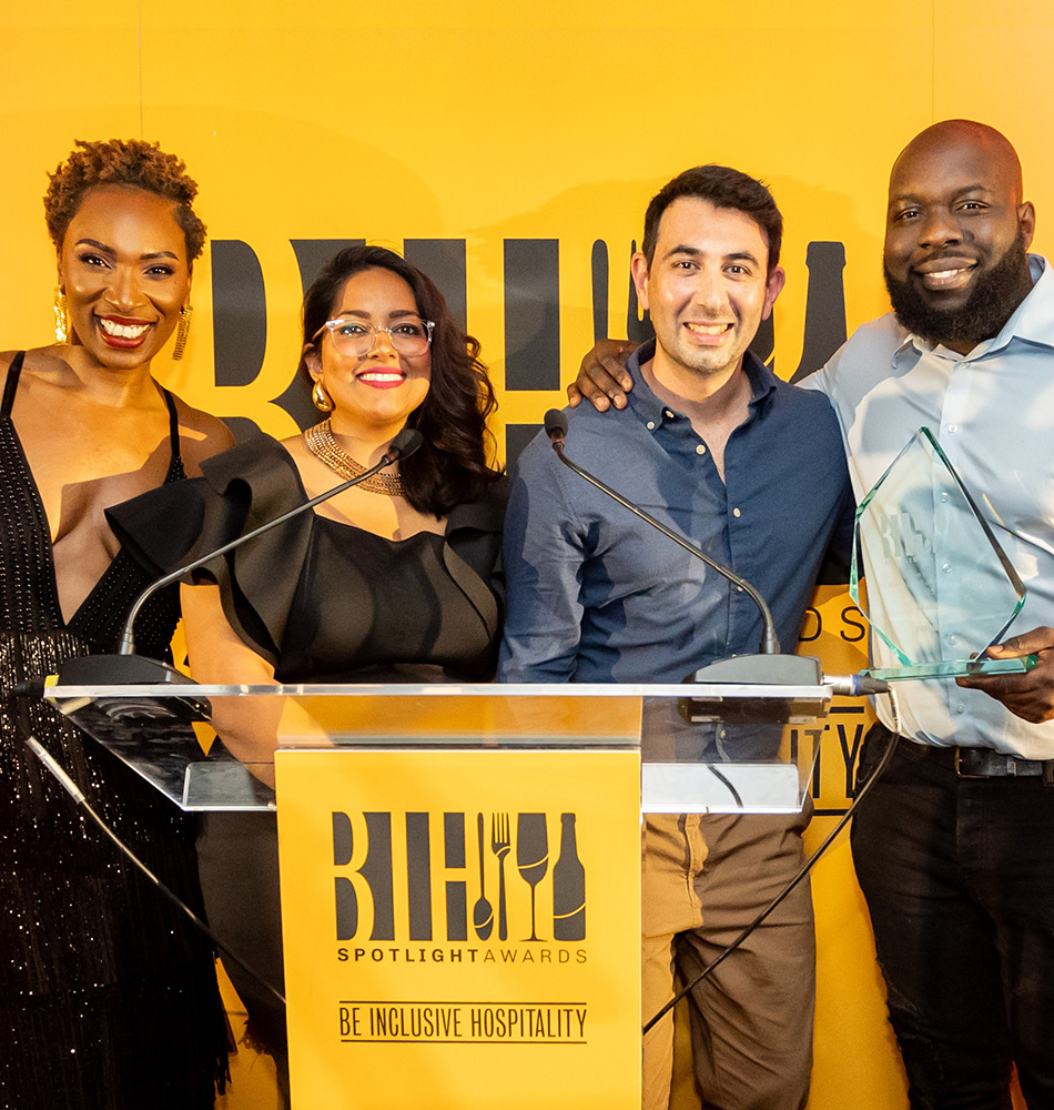 Lorraine Copes and one award recipient, one award host and sponsor, smiling at the Be Inclusive Hospitality Spotlight Awards, standing behind a podium with the event’s logo.