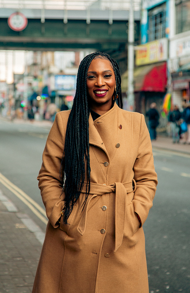 Lorraine Copes standing on a vibrant street, wearing a tan coat, smiling confidently, with Peckham in the background.