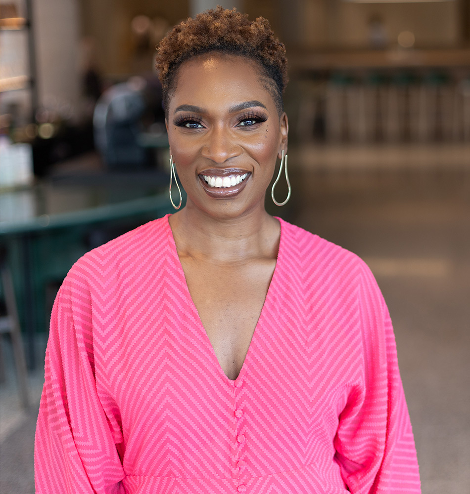 Lorraine Copes smiling, wearing a bright pink dress, with a blurred indoor background, showcasing her friendly and approachable demeanour.