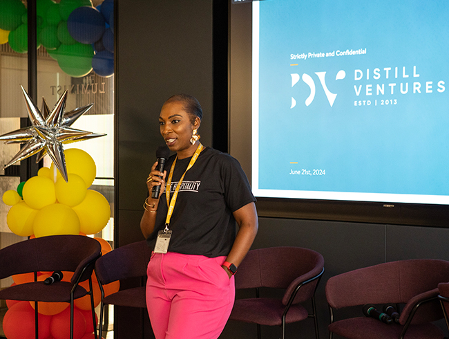 Lorraine Copes speaking at an event, which is a partnership between Distill Ventures and Be Inclusive Hospitality on June 21st, 2024, wearing a Be Inclusive Hospitality T-shirt and pink trousers, standing beside a presentation screen and colourful balloon decorations.