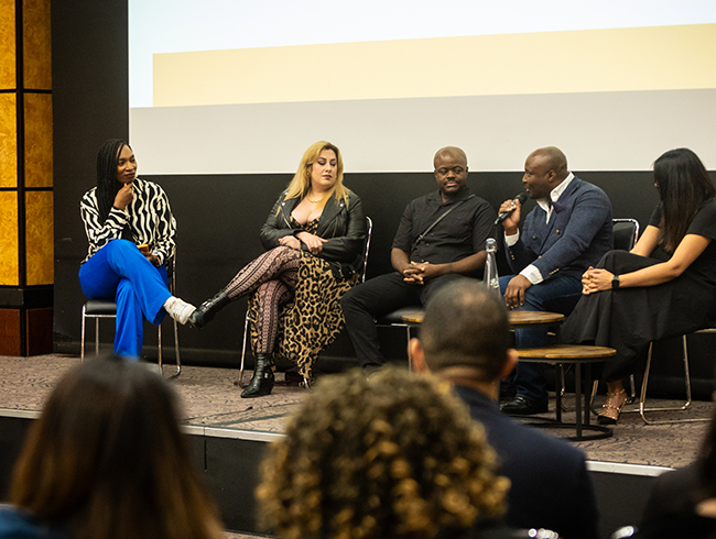 Lorraine Copes chairing a panel discussion with four other hospitality leaders and speakers, seated on stage, engaging with the audience at an event at a Be Inclusive Hospitality community event in February 2024