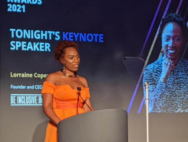 Lorraine Copes delivering a keynote speech at an event in 2021, wearing an orange dress, with ‘Tonight’s Keynote Speaker’ displayed on the screen behind her.